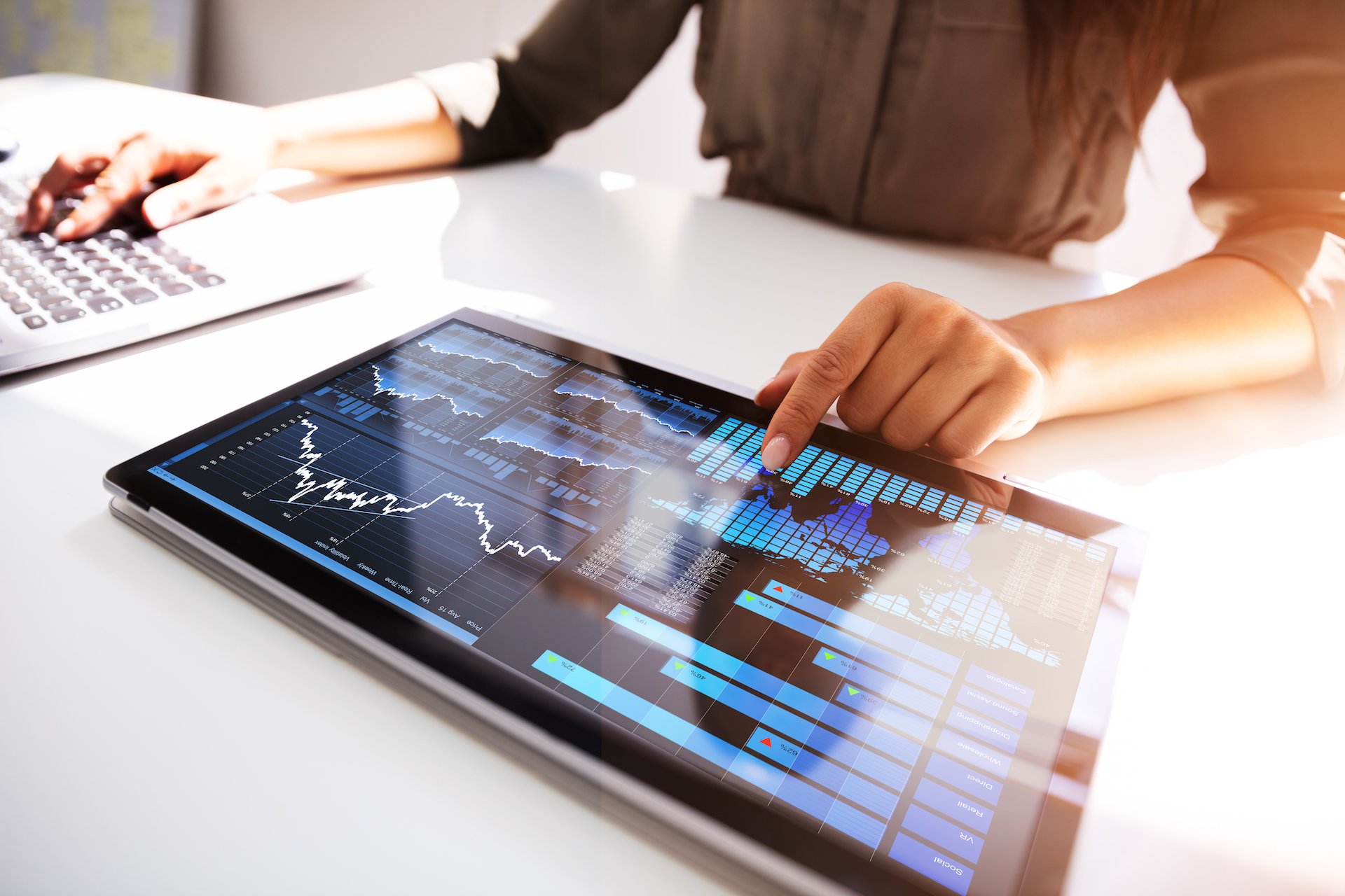 Woman sitting at desk with keyboard and electronic tablet, pointing at digital graphs