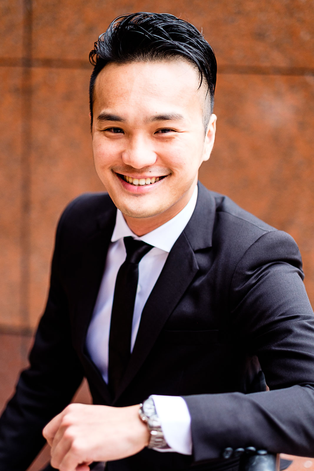 Asian man in black blazer and tie smiling into the camera