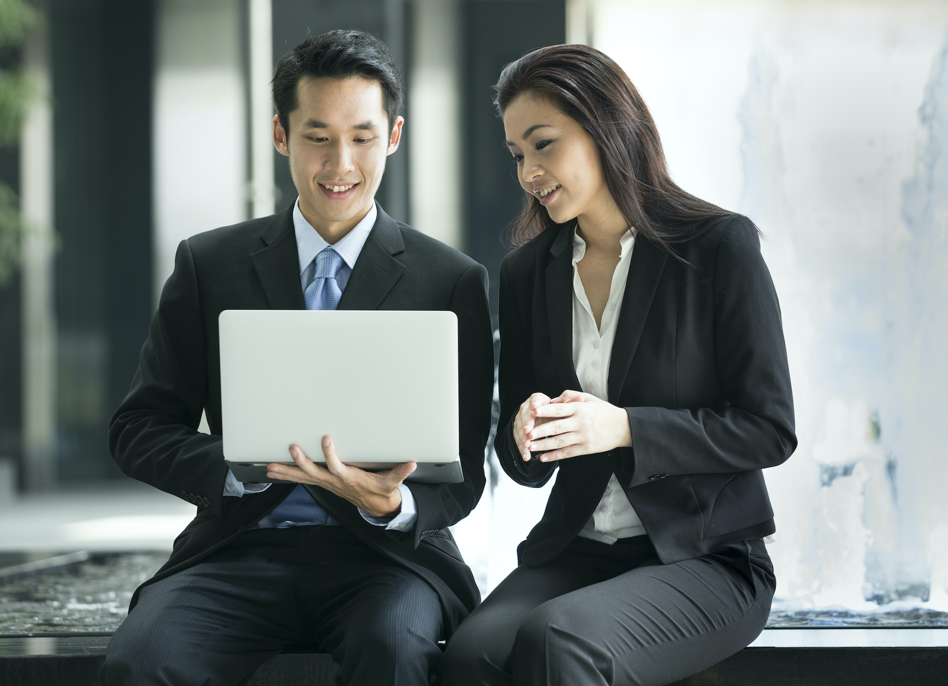 Asian man and woman in dark business suits looking at laptop screen together
